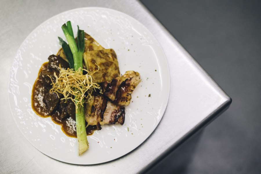 Ein Teller mit Rostbraten, Kartoffelgratin, Lauch und knusprigen Zwiebeln auf einer Metalloberfl&auml;che.