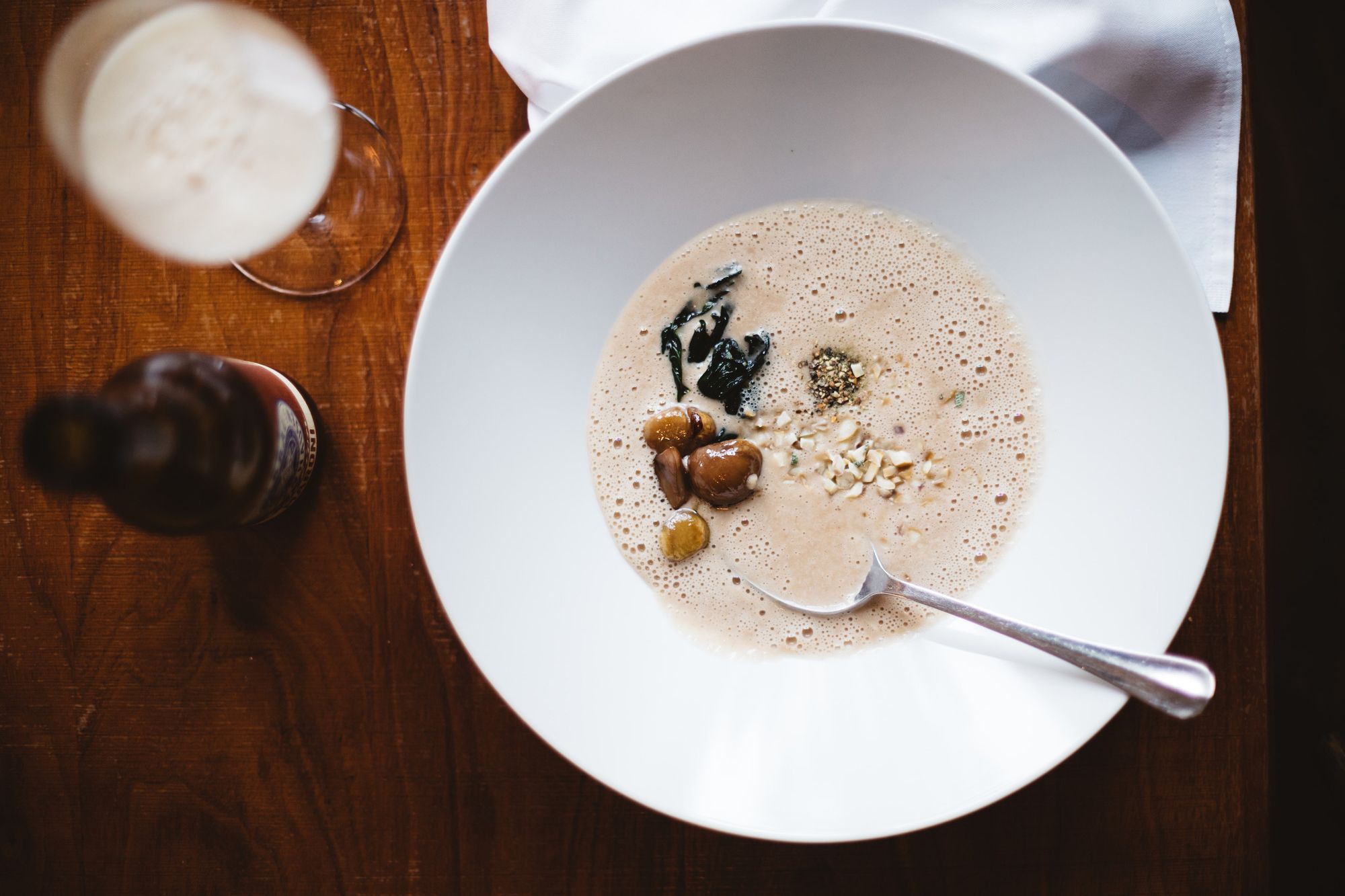 Eine Schüssel Maroni-Suppe mit Nüssen und Kräutern auf einem Holztisch, daneben ein Glas und eine Flasche.