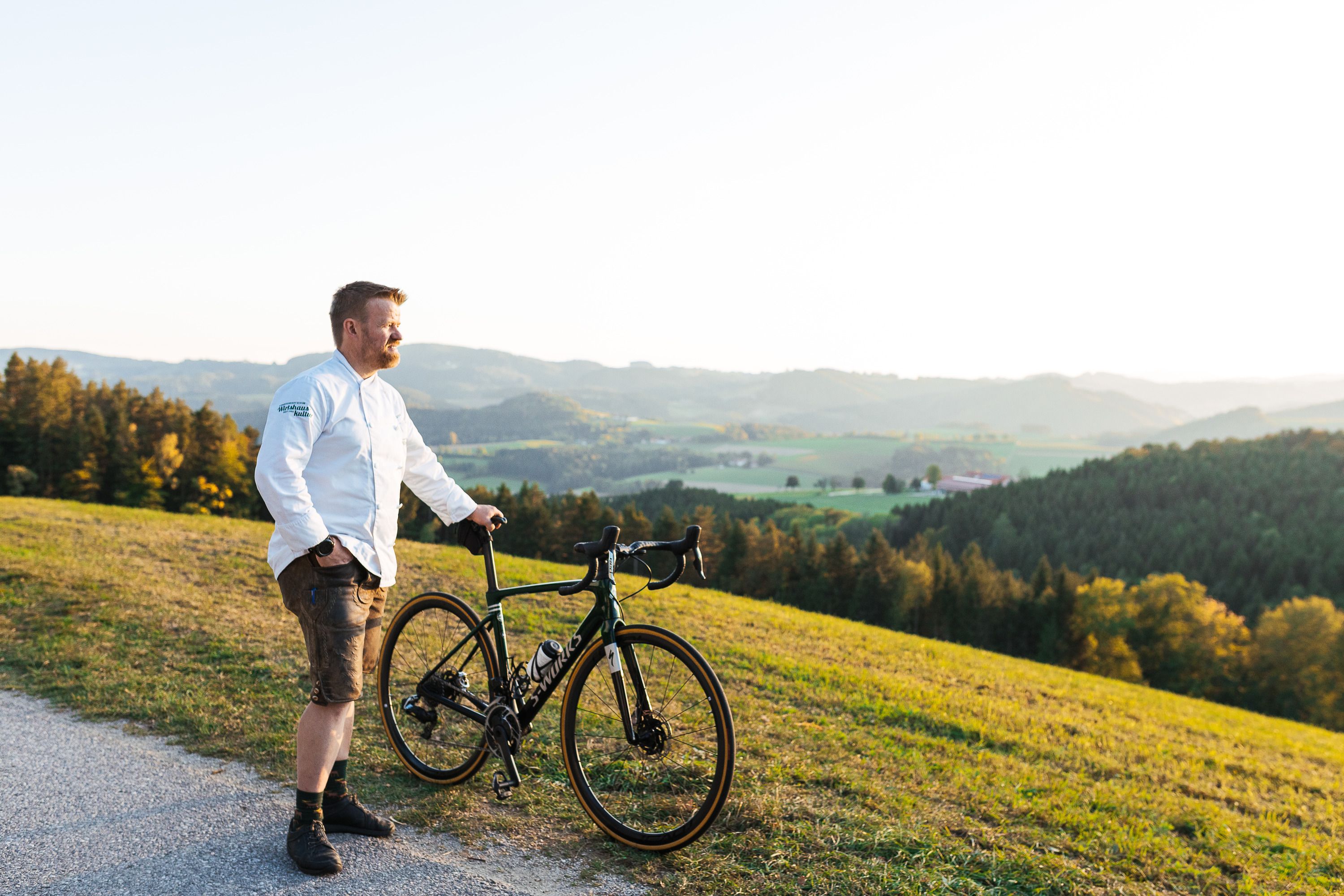 Ein Mann in weißem Hemd steht mit einem Fahrrad auf einem Hügel mit Blick auf eine grüne Landschaft.