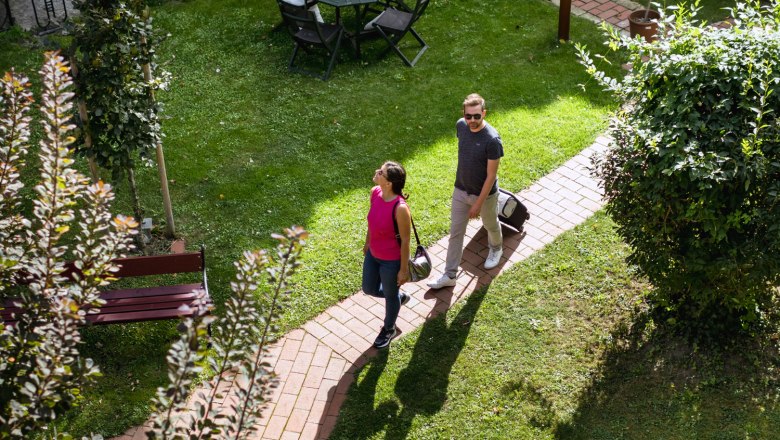 Zwei Personen gehen auf einem gepflasterten Weg durch einen grünen Garten mit Tisch und Stühlen.