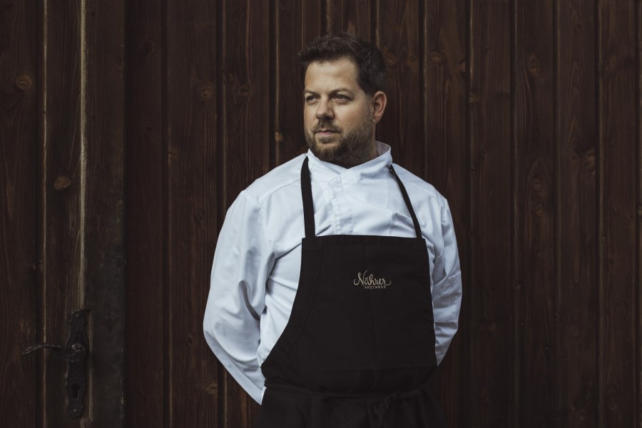 Ein Mann in Kochkleidung steht vor einer Holzwand.