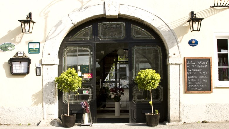 Eingang eines Restaurants mit dekorativen Pflanzen und einer Tafel mit Menüangeboten.