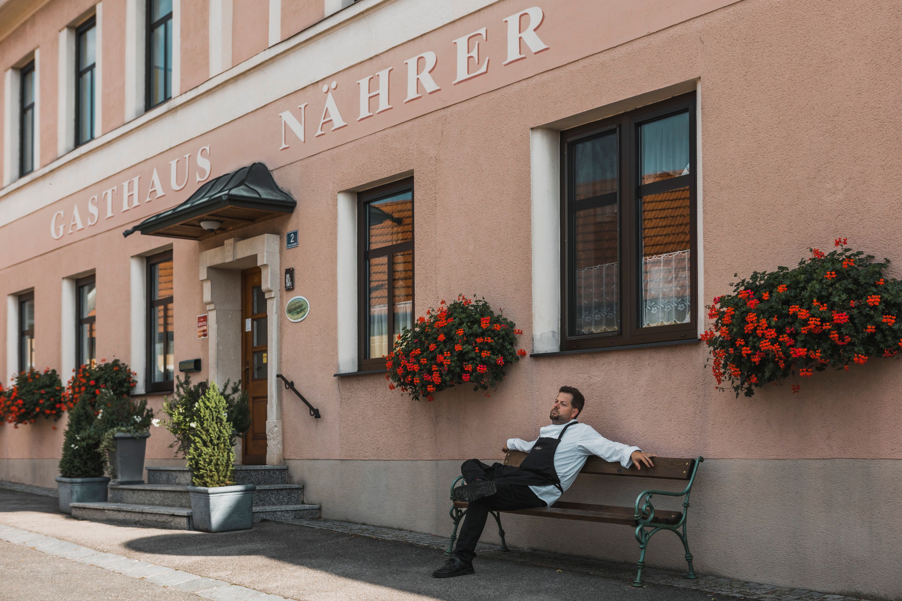 Ein Mann in Kochkleidung sitzt entspannt auf einer Bank vor dem Gasthaus Nährer mit Blumendekoration.