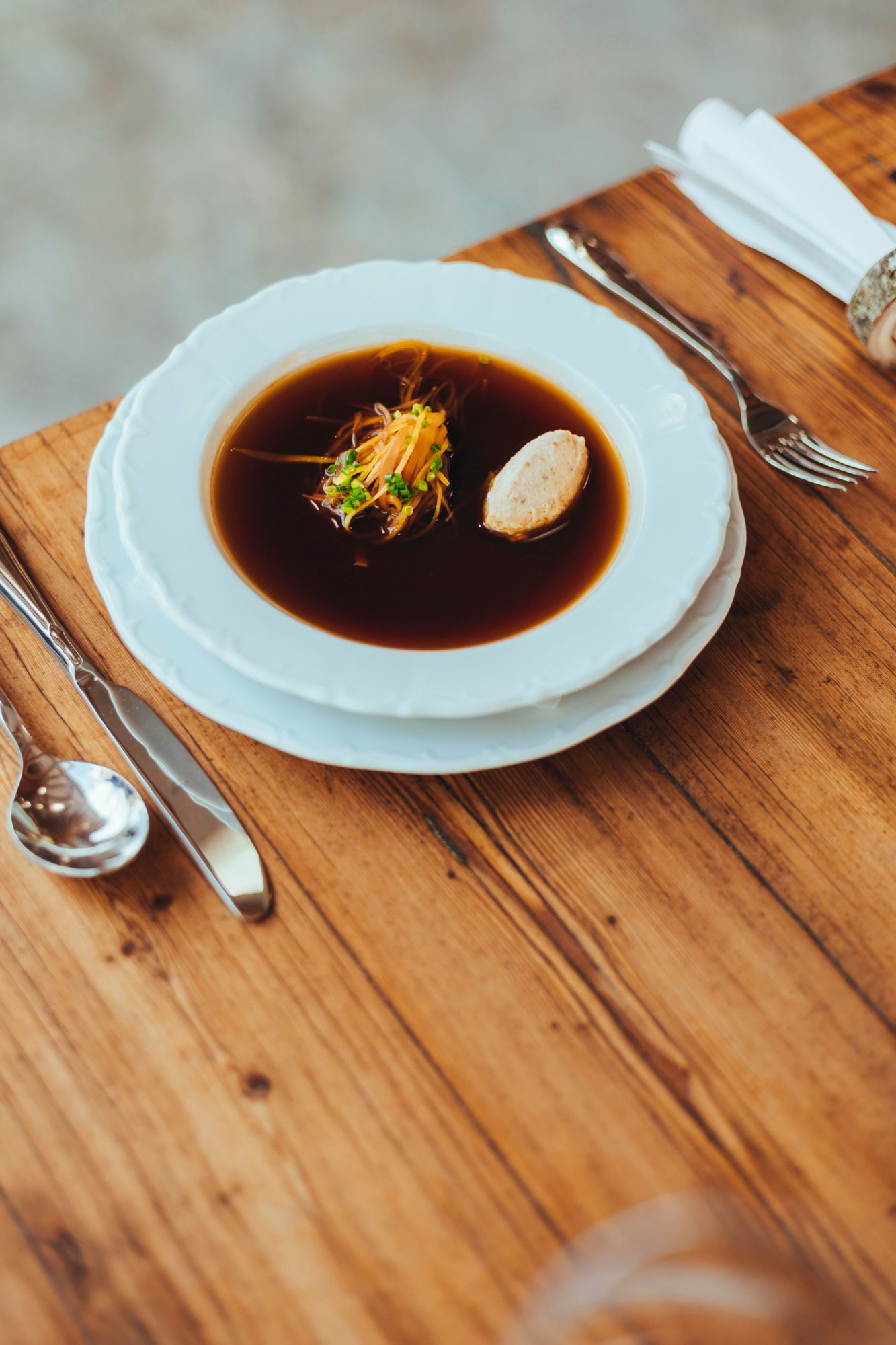 Ein Teller mit klarer Rindssuppe und Einlagen auf einem Holztisch, daneben Besteck.