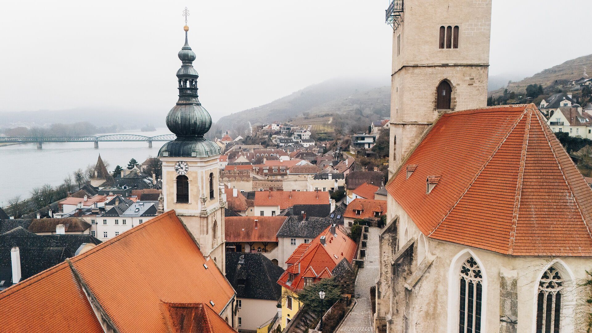 Die sanften Hügel und die charmanten Dächer der Stadt laden zu einem winterlichen Spaziergang ein. Die ruhige Donau spiegelt die grauen Wolken wider und schafft eine friedliche Atmosphäre, die zum Verweilen einlädt.