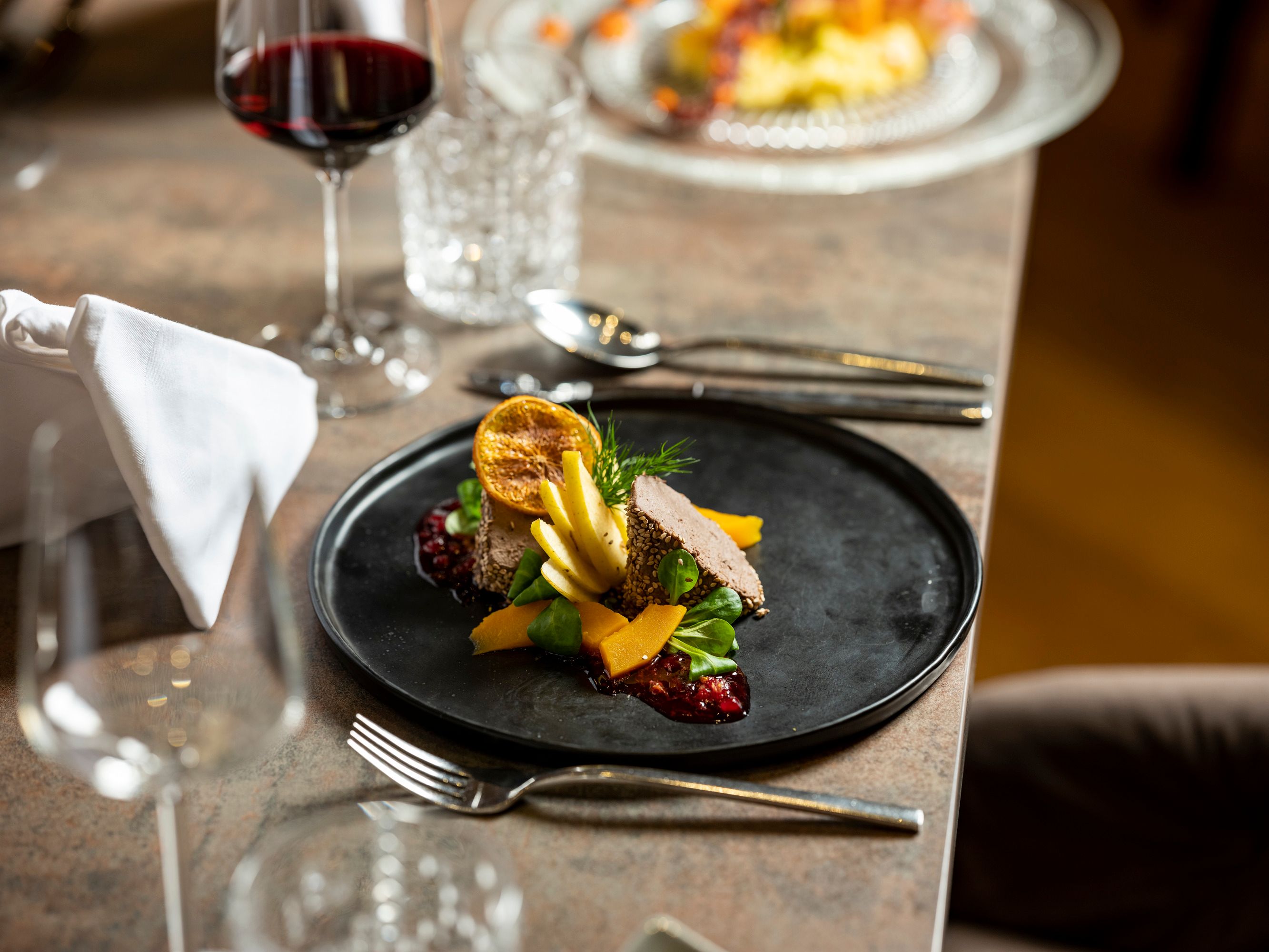 Elegantes Gericht auf schwarzem Teller mit Rotwein im Hintergrund.