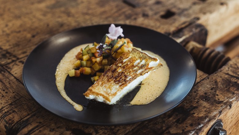 Gebratener Fisch mit Gem&uuml;sew&uuml;rfeln und Sauce auf schwarzem Teller serviert.