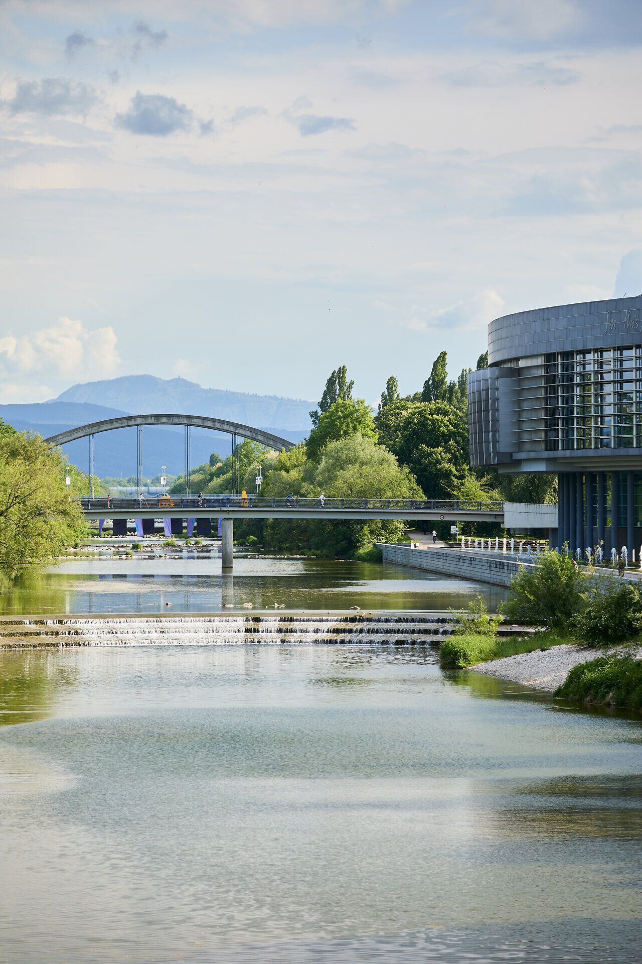Die sanften Wellen des Wassers spiegeln die üppige grüne Landschaft wider, während die Brücke elegant über den Fluss führt. Hier, wo Natur und Architektur harmonisch aufeinandertreffen, lädt die Umgebung zu einem entspannenden Spaziergang ein.