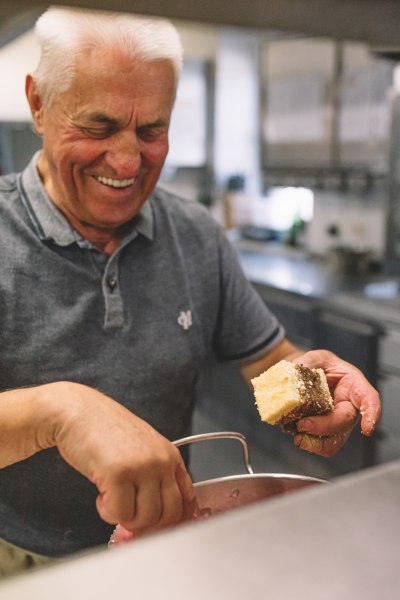Ein &auml;lterer Mann in einer K&uuml;che h&auml;lt ein St&uuml;ck Kuchen und l&auml;chelt.