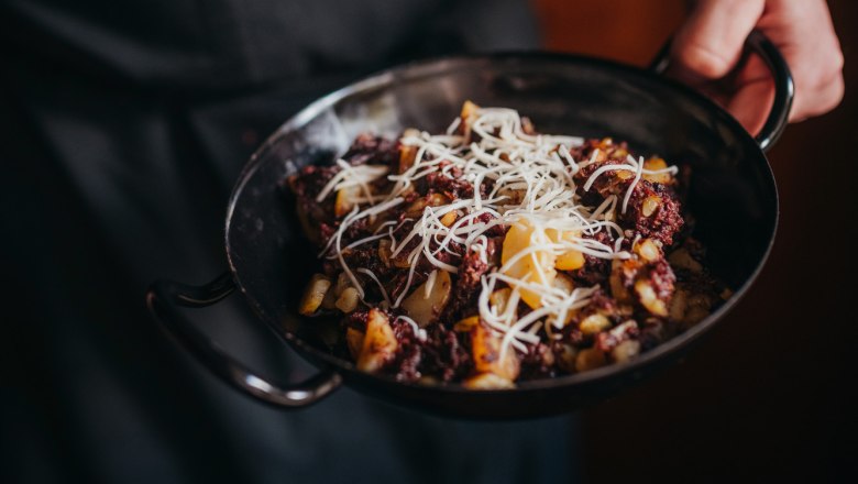 Eine Person h&auml;lt eine Pfanne mit Blunzengr&ouml;stl, garniert mit geriebenem K&auml;se.