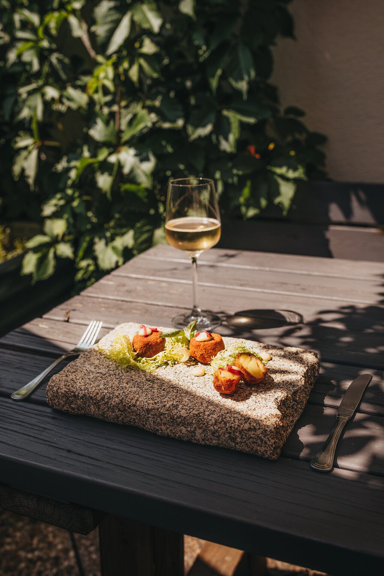 Genießen Sie einen köstlichen gebackenen Erdäpfelsalat, der auf einem rustikalen Stein serviert wird, während ein Glas erfrischender Wein auf dem Tisch steht. Umgeben von üppigem Grün und der warmen Sonne, lädt diese idyllische Atmosphäre zum Verweilen und Schlemmen ein.