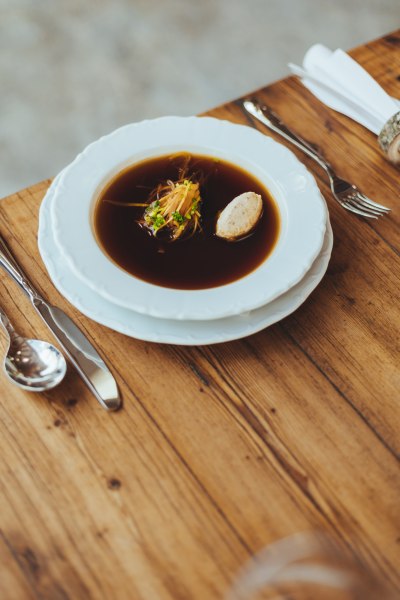 Ein Teller mit klarer Rindssuppe und Einlagen auf einem Holztisch, daneben Besteck.