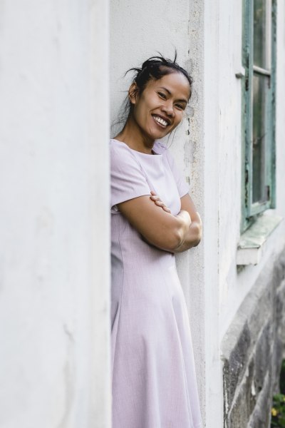 Frau in lila Kleid lehnt lächelnd an einer Wand.