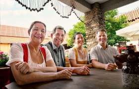 Vier Personen sitzen lächelnd an einem Tisch im Freien, umgeben von Blumen und einem rustikalen Ambiente.
