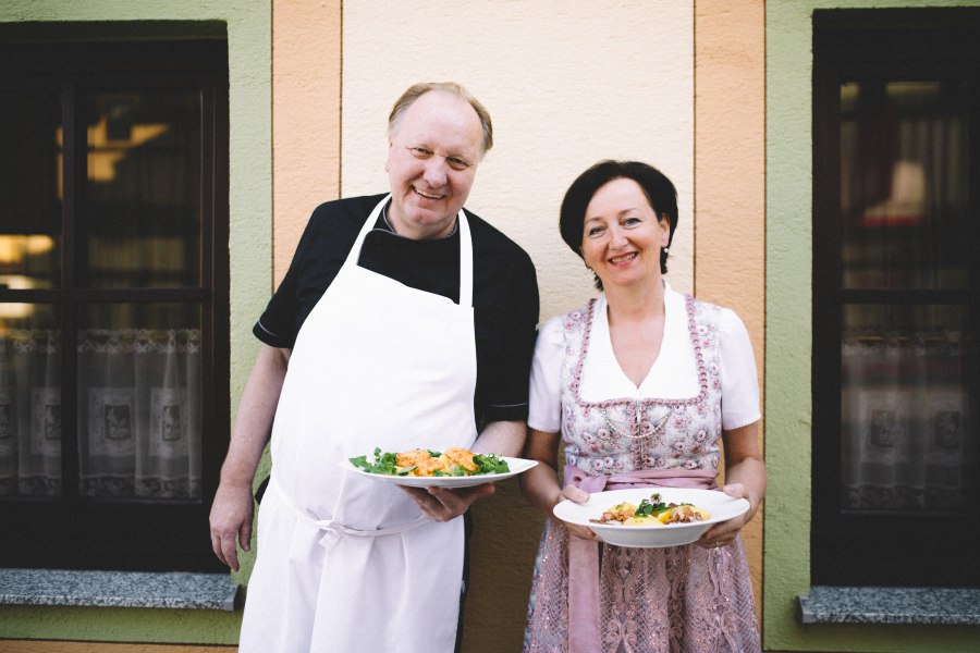 Ein Mann in Sch&uuml;rze und eine Frau in traditioneller Kleidung halten Teller mit Essen vor einem Geb&auml;ude.