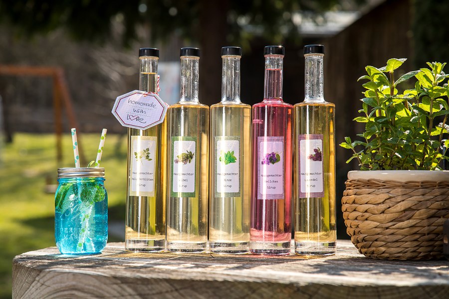 F&uuml;nf Flaschen mit selbstgemachtem Sirup und ein blaues Glas mit Strohhalmen auf einem Holztisch im Freien.