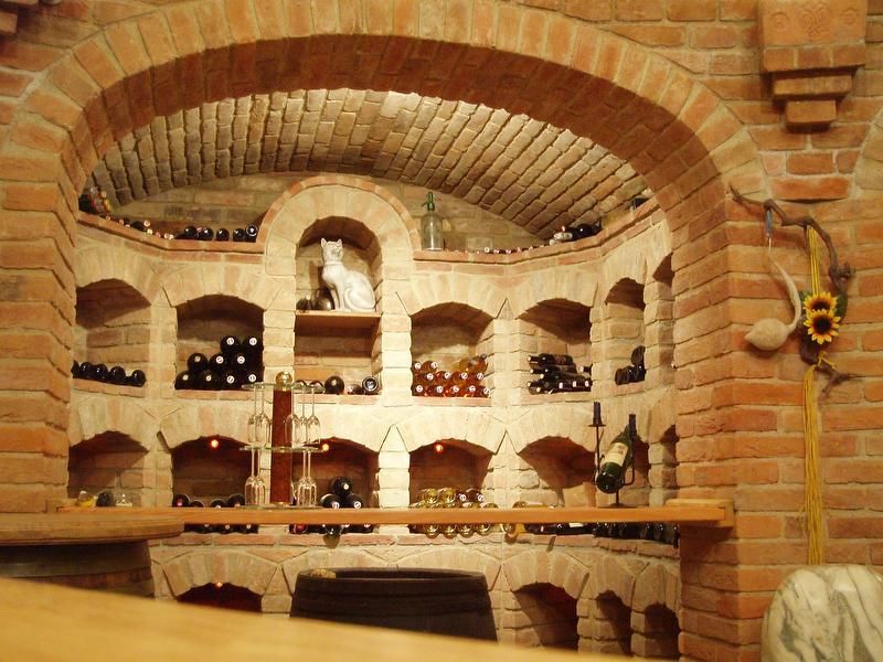 Ein Weinkeller mit gemauerten Regalen, gefüllt mit Weinflaschen. Eine Katzenfigur steht in der Mitte. Eine Sonnenblume hängt an der Wand.