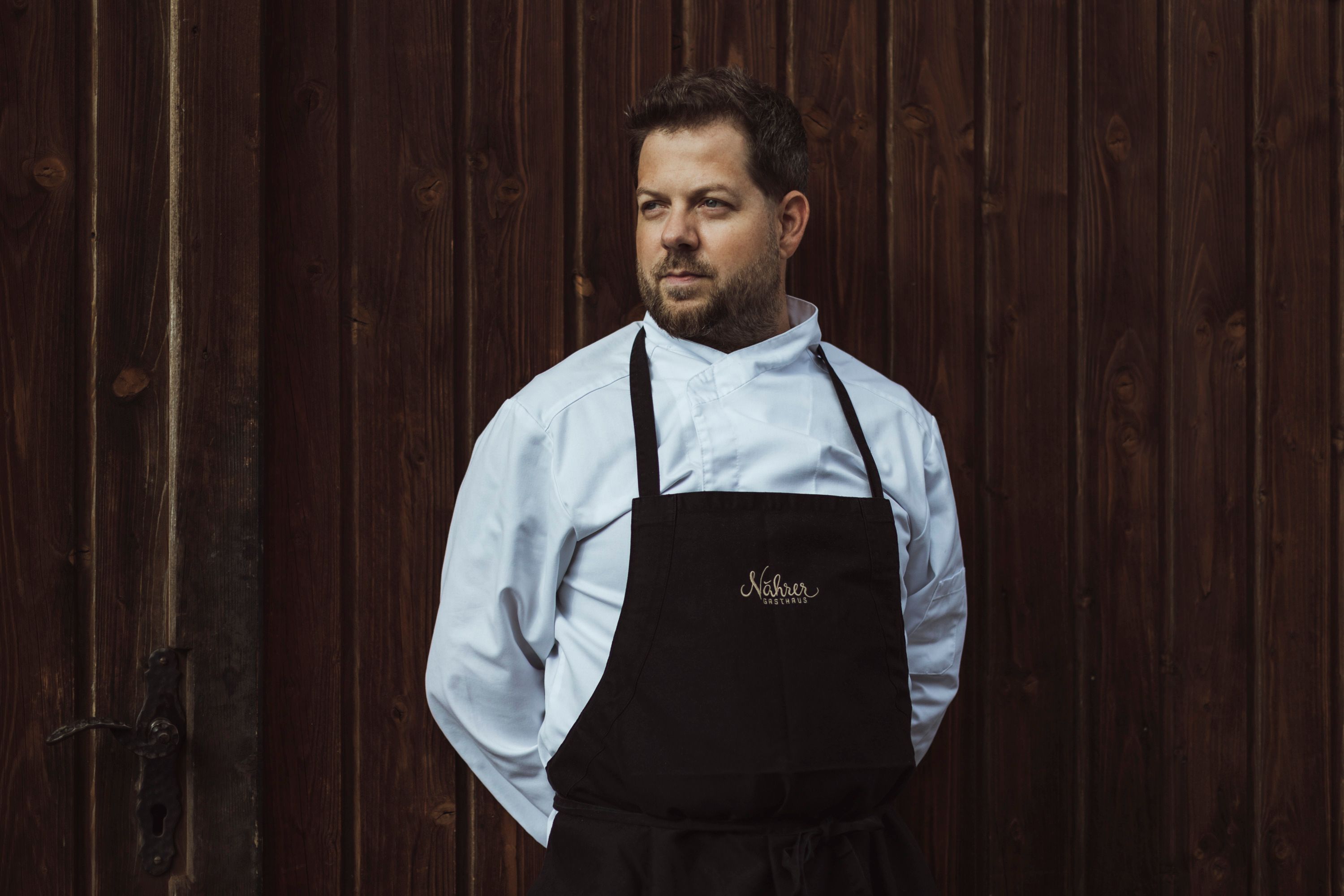 Ein Mann in Kochkleidung steht vor einer Holzwand.