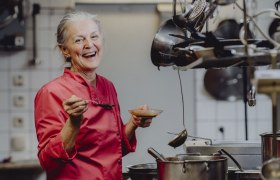 Eine l&auml;chelnde Frau in einer roten Kochjacke steht in einer K&uuml;che mit einem L&ouml;ffel und einer Sch&uuml;ssel in der Hand.