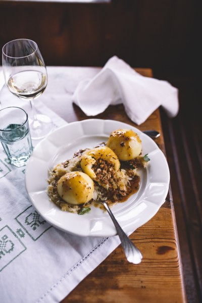 Drei gef&uuml;llte Kn&ouml;del auf Sauerkraut mit So&szlig;e auf einem Teller, daneben ein Glas Wei&szlig;wein.