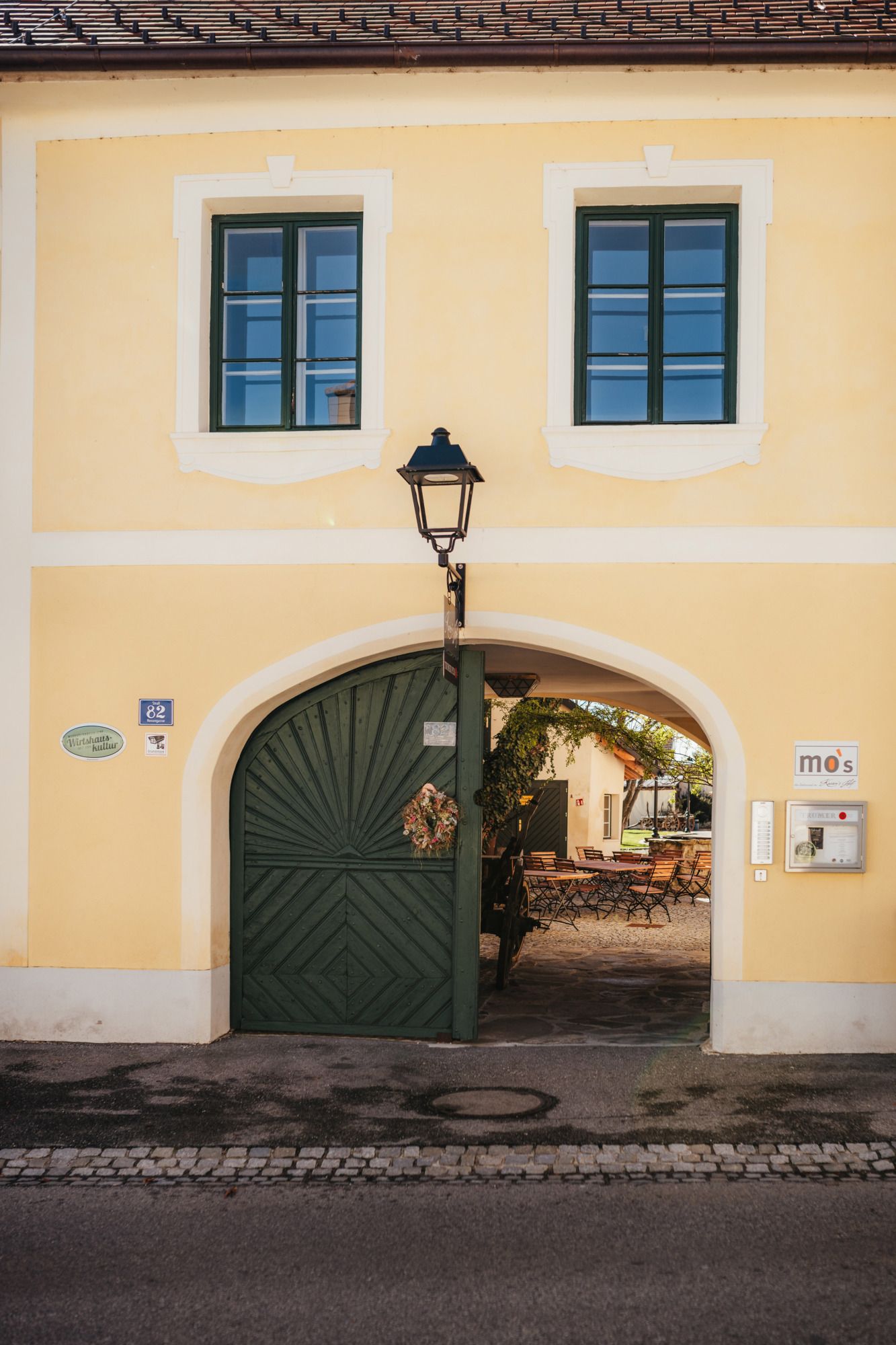 Gelbes Gebäude mit grünem Tor und zwei Fenstern, das zu einem Innenhof führt. Schild mit der Aufschrift 'mo's'.