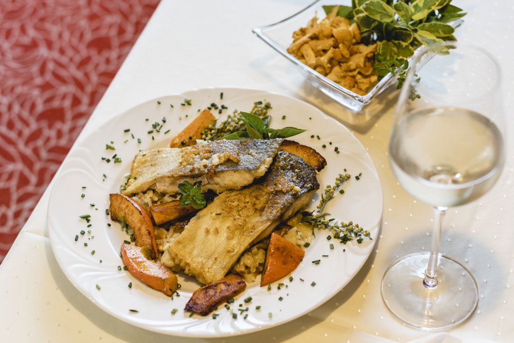 Gourmetgericht mit Fischfilet, Gemüse und Weißwein auf weißem Tisch.