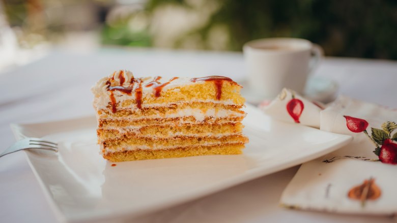 Ein St&uuml;ck Schichttorte mit Sahne und Karamell auf einem wei&szlig;en Teller, daneben eine Tasse Kaffee und eine Serviette mit Fruchtmuster.