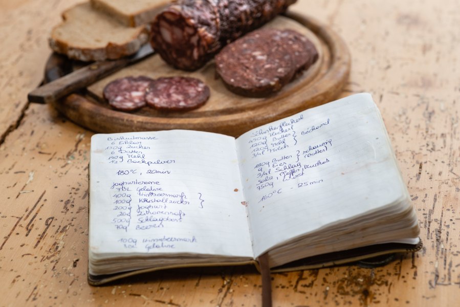 Handgeschriebenes Rezeptbuch liegt auf einem Tisch neben einer Holzplatte mit Wildwurst und Brot.
