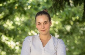 Eine Frau in weißem Hemd steht mit verschränkten Armen vor einem unscharfen, grünen Hintergrund.