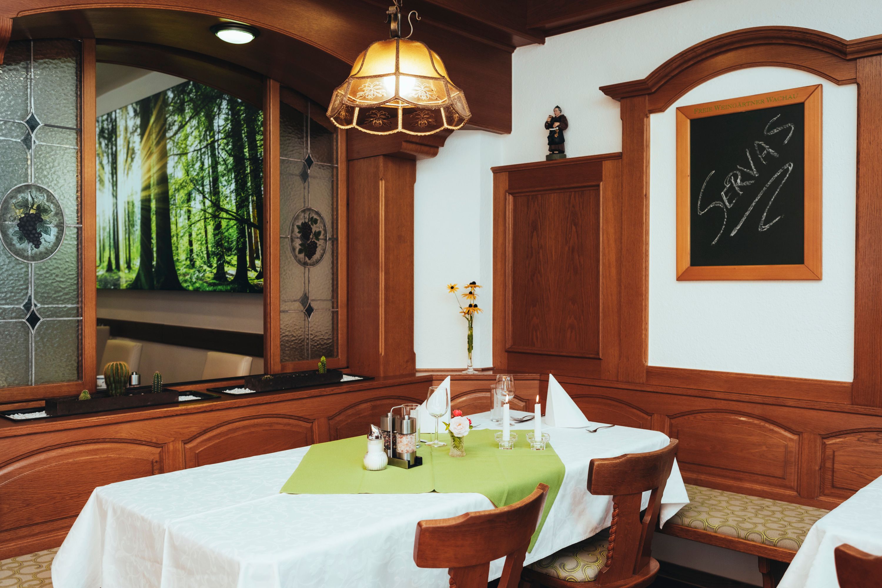 Gemütlicher Gastraum mit Holzmöbeln, Tisch mit grüner Tischdecke, Kerzen und Dekoration. Fenster mit Waldmotiv und Tafel mit Schriftzug 'Servas'.