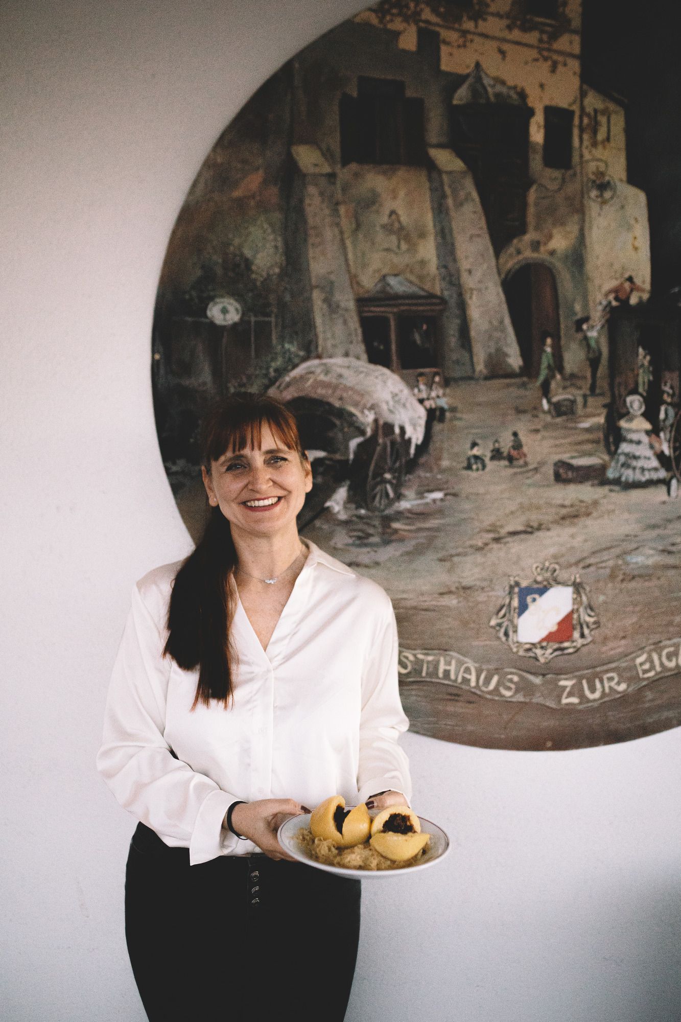 Frau in weißer Bluse hält Teller mit Knödeln vor einem Wandgemälde.