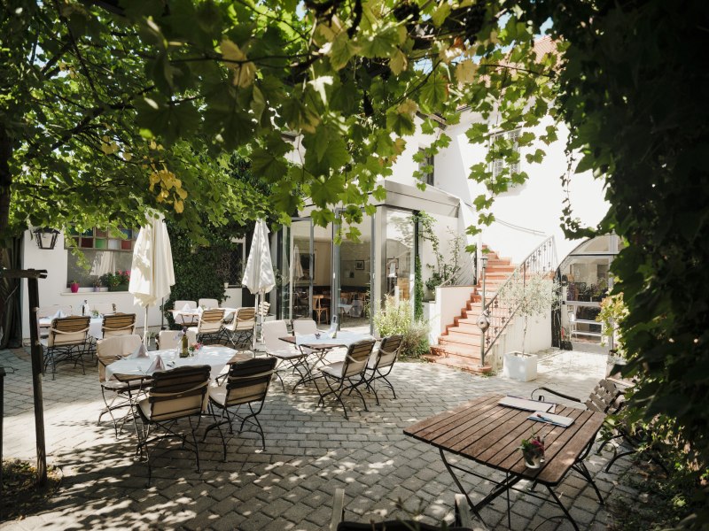 Gem&uuml;tlicher Innenhof mit Tischen und St&uuml;hlen unter gr&uuml;nen Bl&auml;ttern.