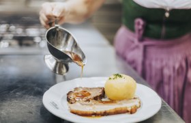 Eine Person gießt Soße über einen Teller mit Braten und Knödel.