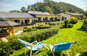 Zwei blaue Liegest&uuml;hle auf einer gr&uuml;nen Wiese vor einem gelben Geb&auml;ude mit Blumen auf den Balkonen.