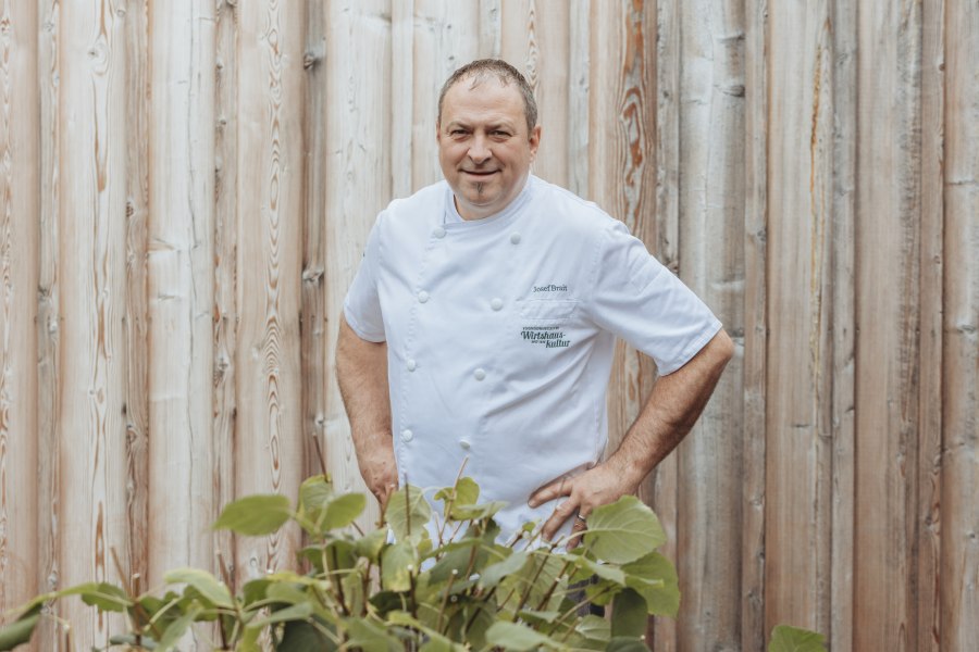 Ein Mann in wei&szlig;er Kochjacke steht vor einer Holzwand.