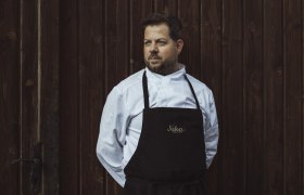 Ein Mann in Kochkleidung steht vor einer Holzwand.