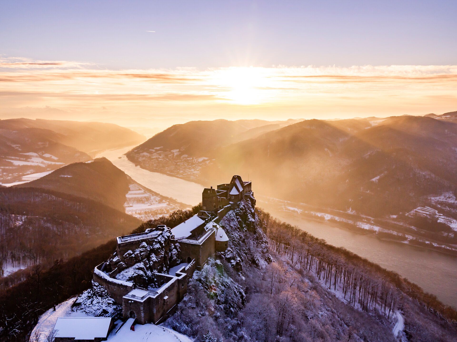 Die schneebedeckte Burgruine Aggstein im Winter mit dem Donautal un der untergehenden Sonne im Hintergrund.