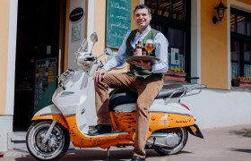 Ein Mann sitzt auf einem Roller vor einem Geb&auml;ude und h&auml;lt ein Tablett mit Bierflaschen und einem Glas.