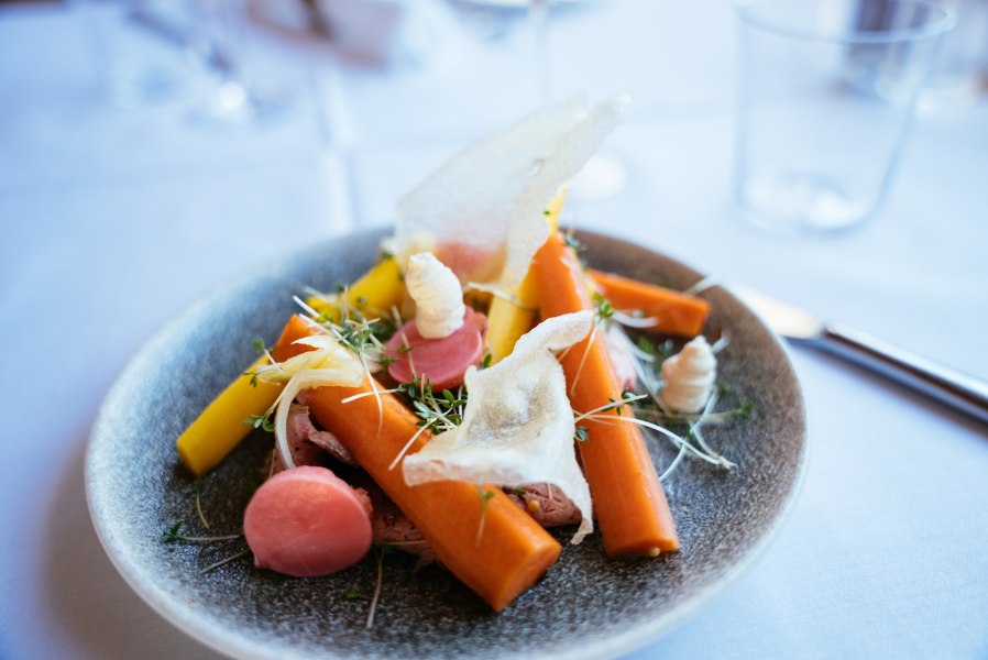 Ein kunstvoll angerichtetes Gericht mit Karotten, Radieschen und Kr&auml;utern auf einem grauen Teller.