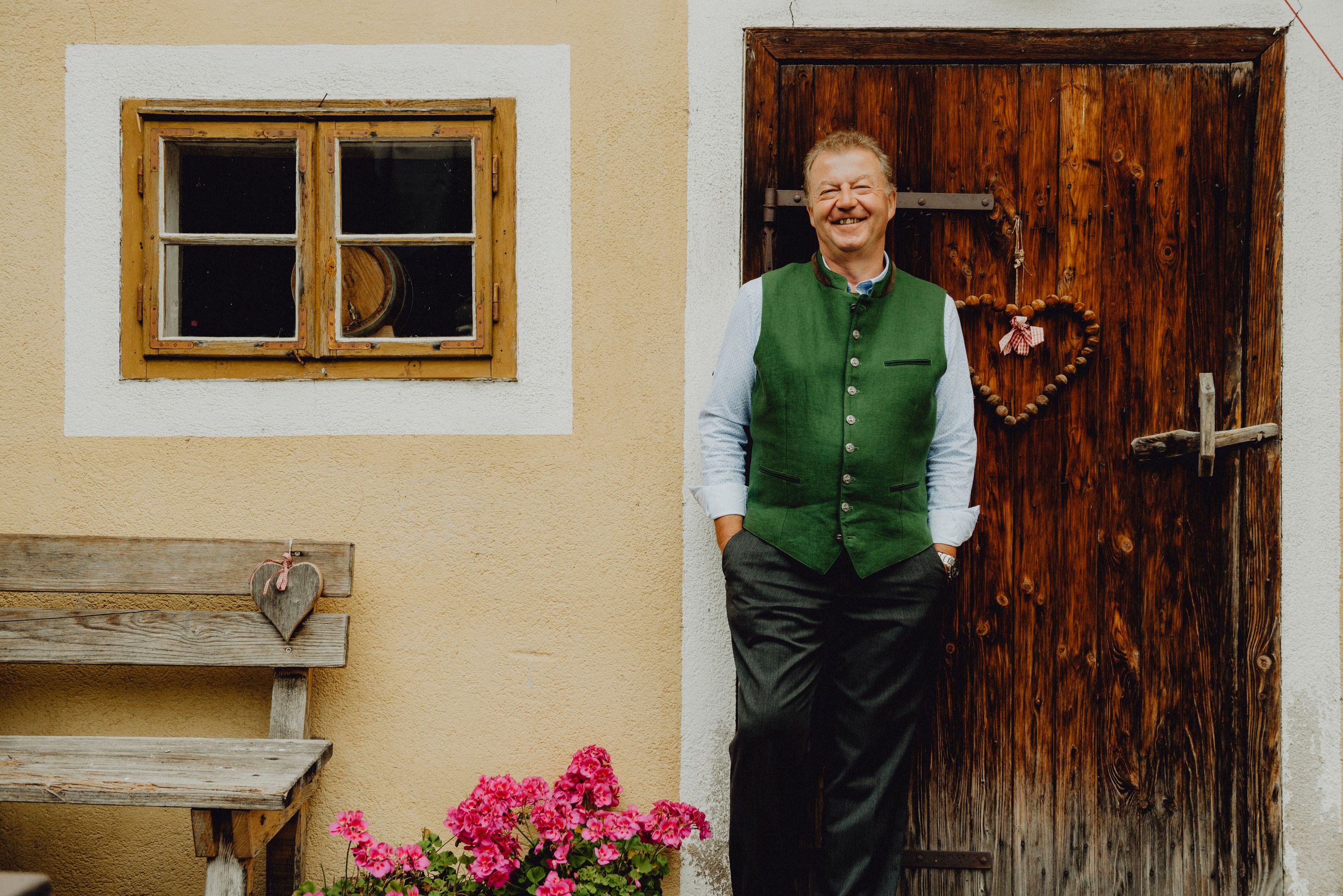 Ein Mann in traditioneller Kleidung steht lächelnd vor einer Holztür neben einem Fenster und einer Bank mit Blumen.