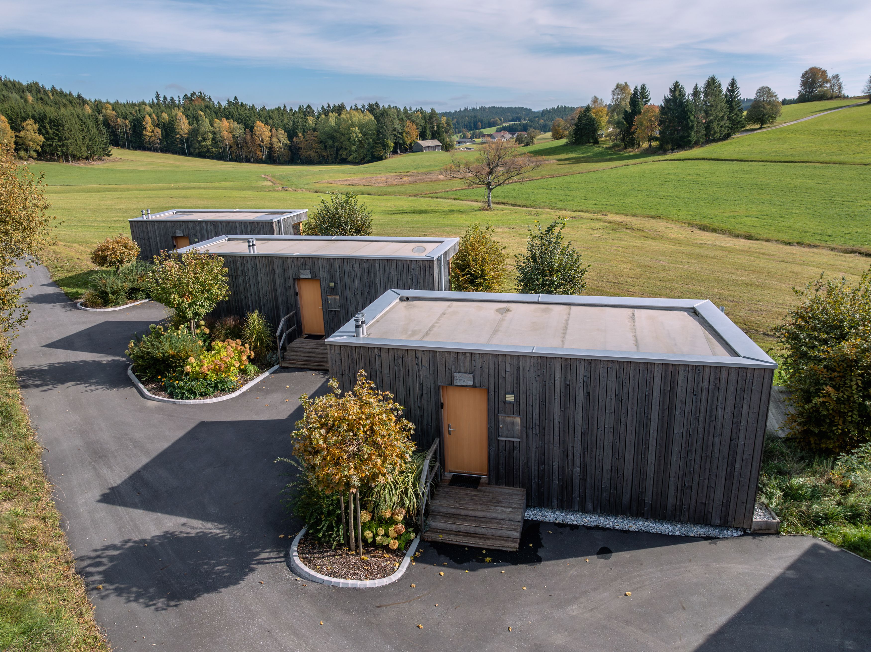 Drei moderne Holzhäuser in einer grünen Landschaft mit Bäumen und Wiesen.