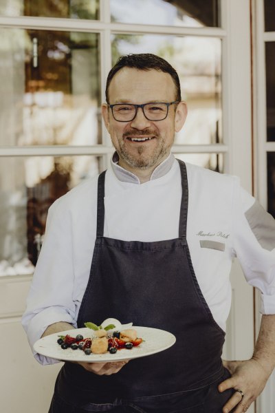 Ein Koch in weißer Uniform und schwarzer Schürze hält einen Teller mit Dessert und Beeren.