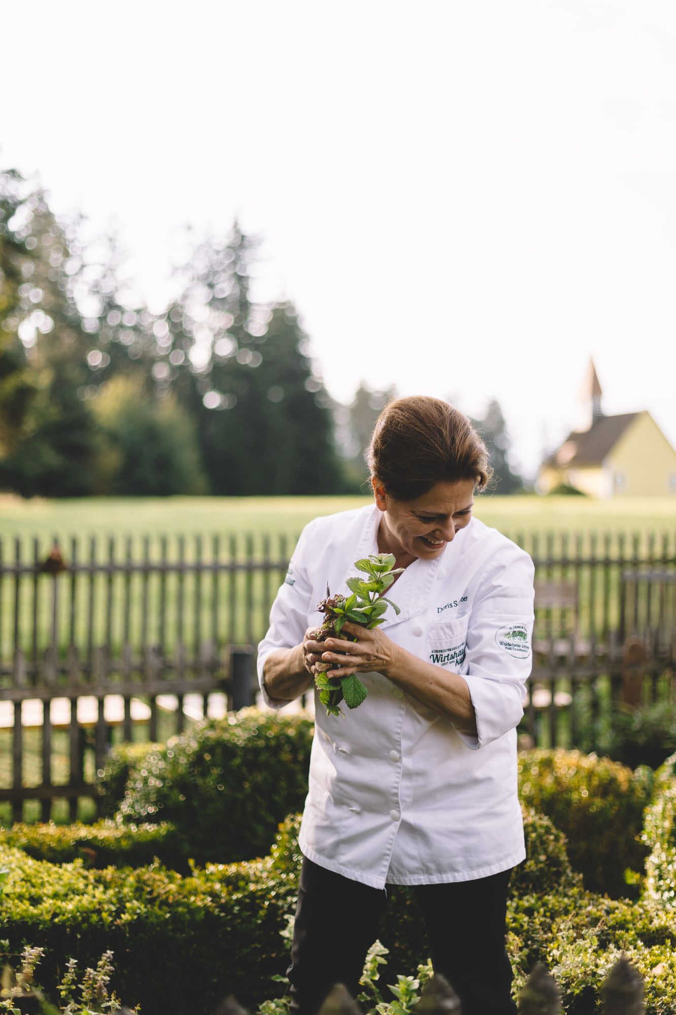 Eine Köchin in weißer Uniform pflückt Kräuter in einem Garten.