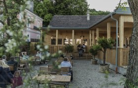 Ein gem&uuml;tlicher Biergarten mit Holzm&ouml;beln und einem Geb&auml;ude im Hintergrund.