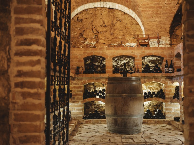 Ein rustikaler Weinkeller mit Ziegelw&auml;nden, Weinregalen und einem Holzfass in der Mitte.