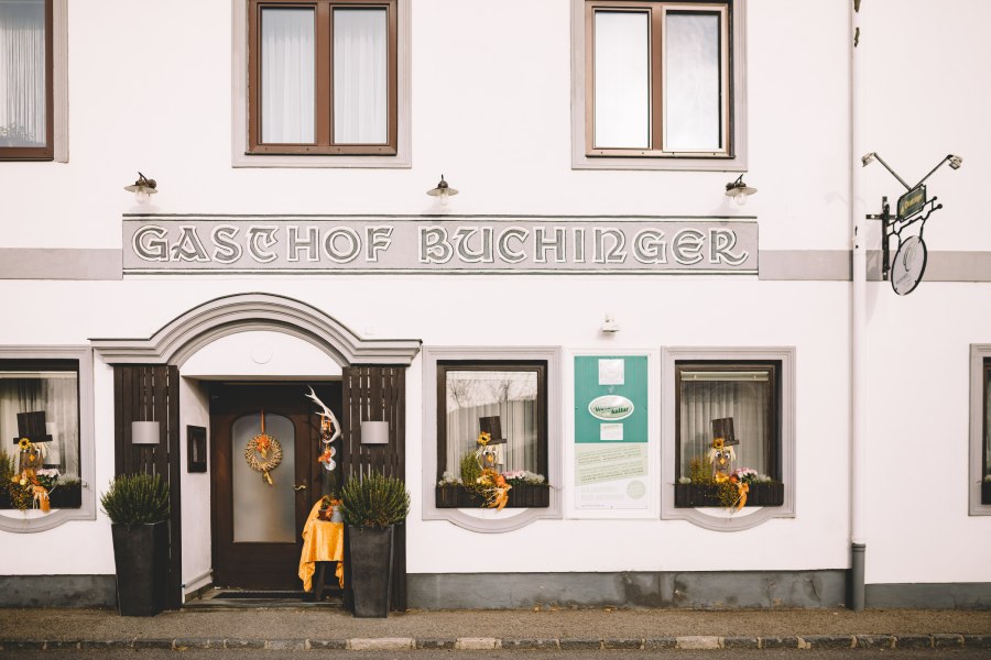 Fassade eines Gasthofs mit der Aufschrift 'Gasthof Buchinger'. Dekorationen und Pflanzen schmücken den Eingang.