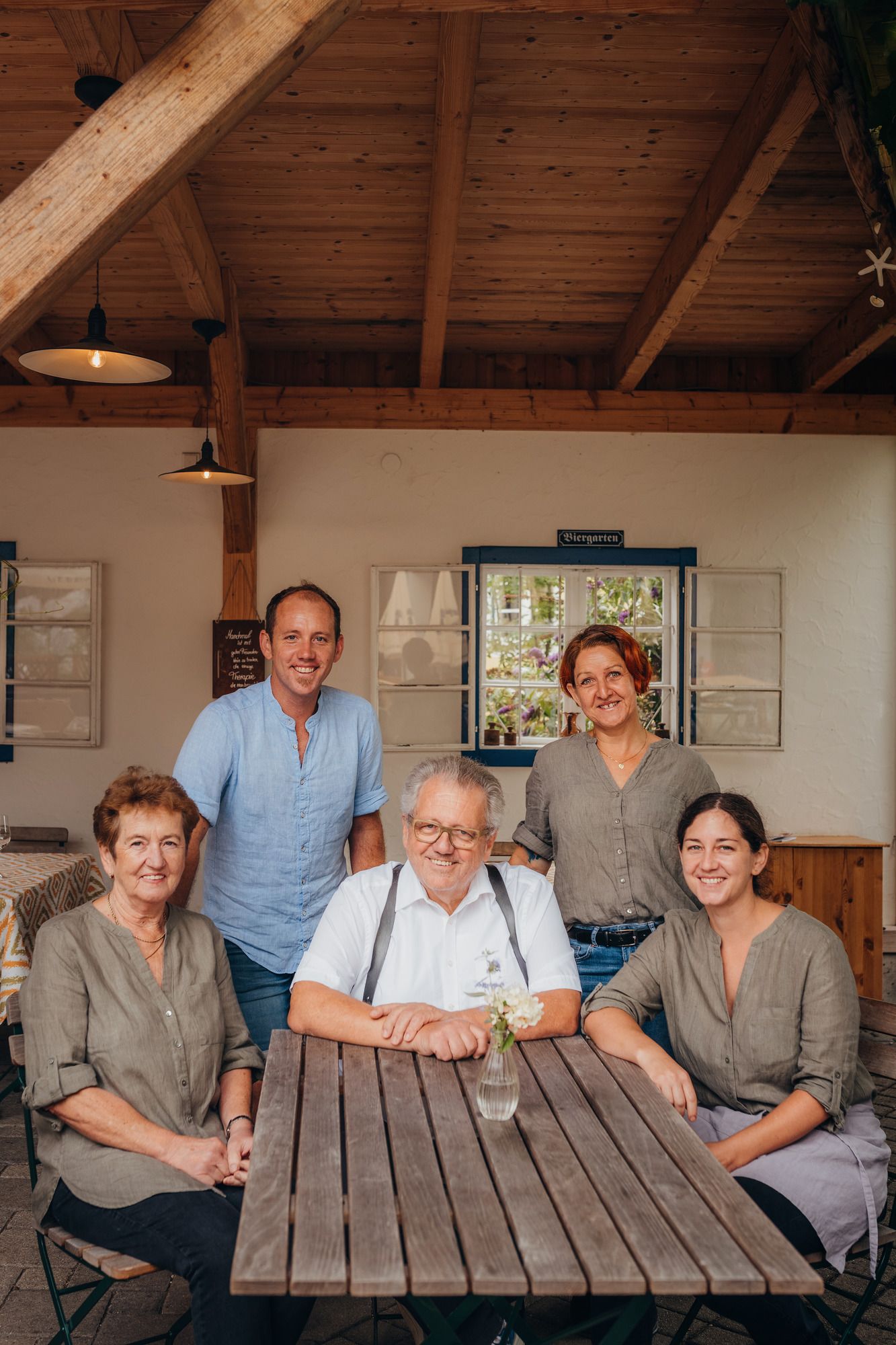 Five people sit and stand around a wooden table in a covered outdoor area.