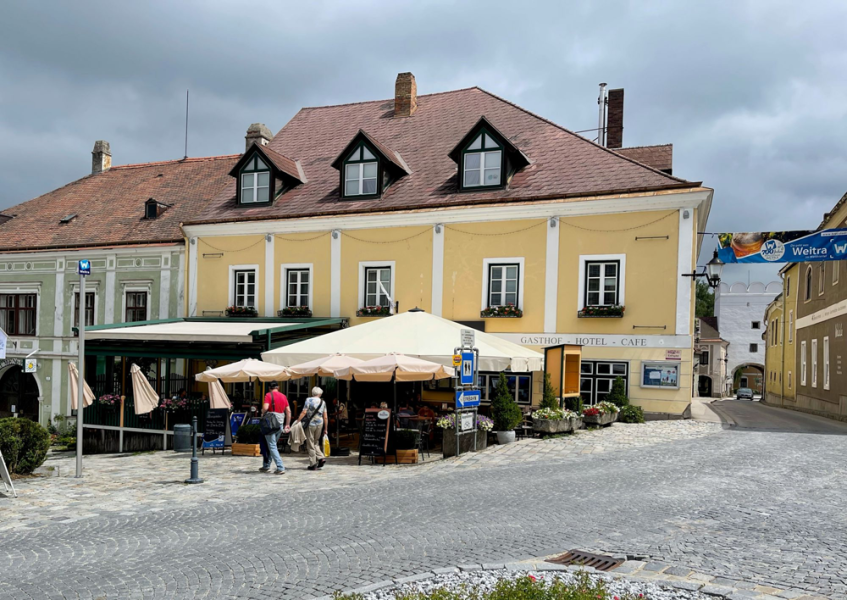Ein gelbes Gebäude mit der Aufschrift 'Gasthof Hotel Cafe' in Weitra, Österreich. Vor dem Gebäude sind Tische mit Sonnenschirmen und Passanten zu sehen.