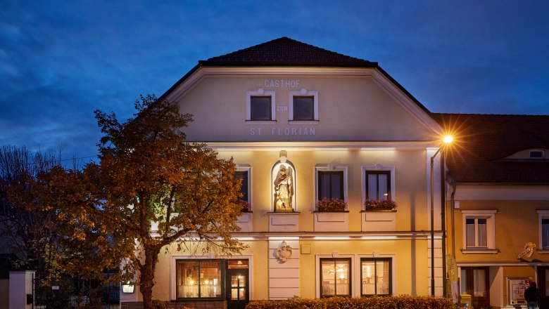 Gasthaus St. Florian bei Dämmerung mit beleuchteter Fassade und Statue.