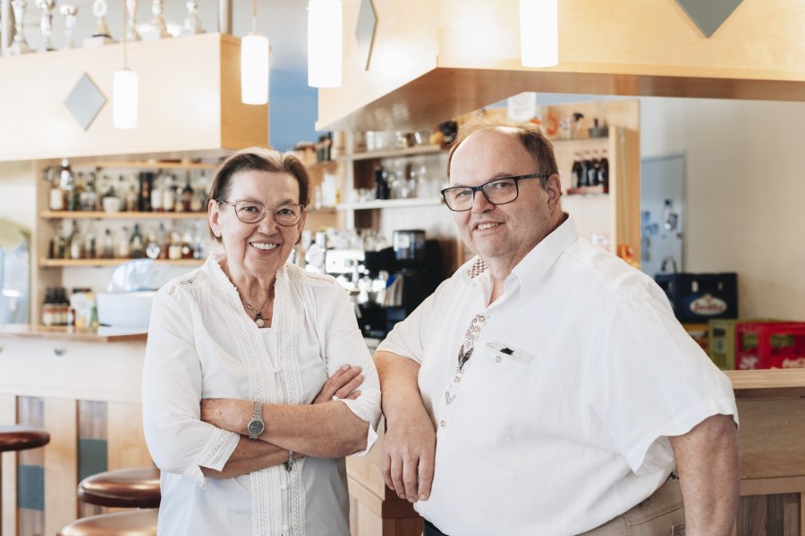 Zwei Personen in einem Caf&eacute; oder einer Bar, l&auml;chelnd, vor einer Theke mit Flaschen im Hintergrund.